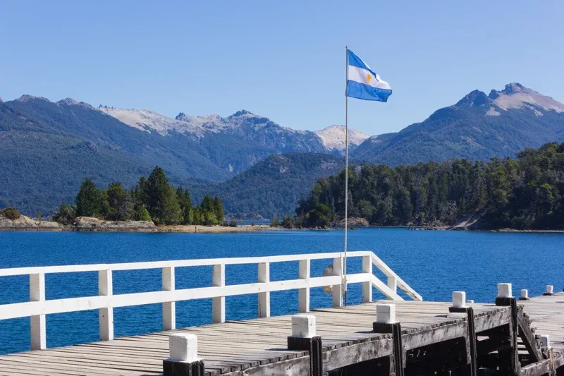 The Argentine flag flies on a dock by a lake, with mountains in the background