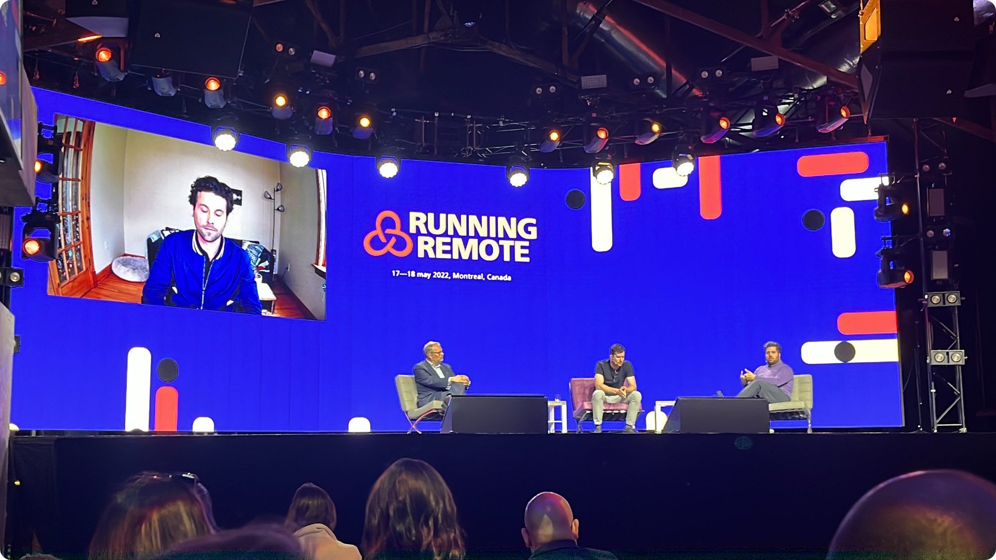 Panelists sit on a Running Remote stage, flanked by a virtual attendee in a conference venue