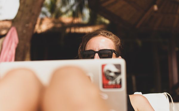 A remote worker on a workation wearing sunglasses works on their laptop (which has a sticker on it)